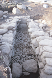 細長い通路の両側に複数の大きな石が積み上げられ、その通路の底には小さな石が敷き詰められている、桃木原古墳群第1号墳とされる遺跡の発掘現場の写真