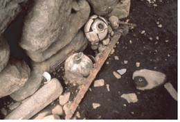 泥の地面に壊れた陶器の壺や錆びた鉄の棒、複数の石が散乱している、古墳の内部から副葬品が発掘された瞬間の写真