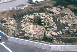 道路脇の柵で囲まれた発掘現場に、石が円形に積まれ中心に入口のような穴がある遺跡の発掘調査を上空から捉えた写真