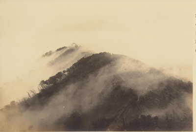 朝霧が立ち込める山間の風景で、前景の山の稜線には樹木が生い茂り、遠景にはさらに大きな山が霞んで見えるセピア色の写真