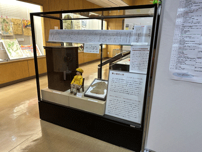 展示室の一角で、ガラスケースの中に座っている黄色い服と帽子を着た「青い目の人形」と、その傍らに展示されている人形の着物や解説パネルを写した写真