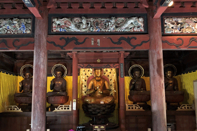 柱と梁で構成された仏堂内部、中央に金色の仏像が安置され、その両脇に2体ずつ茶色の仏像が配置され、上部に彩色彫刻欄間と波模様装飾が施された寺院本堂の写真