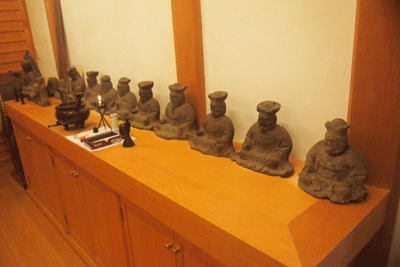 木製の棚の上に灰色の石像が複数並び、中央に燭台とお供え物、香炉が置かれ、白い壁と木製の柱が背景にある仏教寺院の内部の写真