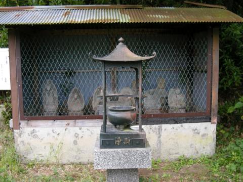 トタン屋根の小屋の中の金網越しに並ぶ、石造六地蔵尊像が6体が横に並んでおり、その手前に設置された石台に香炉が置かれている写真