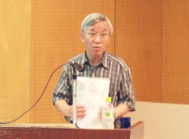 茶色いストライプのシャツを着た白髪の清水先生が、講演台の前に立ち、資料を手に持ち話をしている様子の写真
