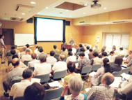 会場で、大勢の参加者たちが椅子に座って前方にあるスクリーンと清水先生に注目している、プロジェクターを使った講演会の様子の写真