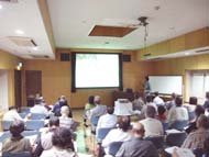 広い会議室で行われた講演会で、多数の参加者たちが椅子に座ってスクリーンを見ている様子の写真