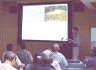 薄暗い室内で、大勢の参加者が椅子に座り、壇上の講師がスクリーンの横で話をしている様子の写真