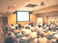 多数の参加者が椅子に座り、壇上の大倉さんがスクリーンを指し示しながら熱心に説明している講演会の様子の写真