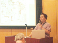 講師の大倉さんが、演壇で片手にマイクを持ち、プロジェクターで映し出された資料を背景に講演している様子の写真