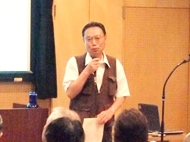 マイクを片手に、白いシャツと茶色いベストを身につけた櫛田さんが、聴衆に向かって熱心に話している様子の写真