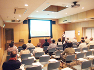 多数の参加者が椅子に座り、壇上の櫛田さんがスクリーンを見ながら説明している講演会の様子の写真