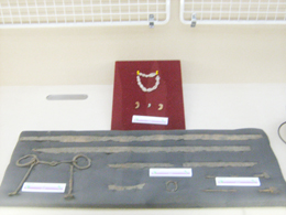 グレーの台の上に複数の金属製の展示物が並び、奥には赤いパネルに白い勾玉のような形の装飾品が展示されている写真