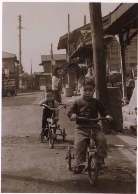 2人の男のが両側に家が立ち並んだ通りを三輪車で走っている古いモノクロ写真