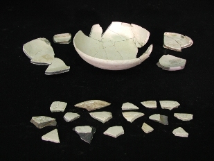 破片をつないで修復した、部分的に欠損がある白い陶器の器の周りに、多数の小さな破片が並べられている写真