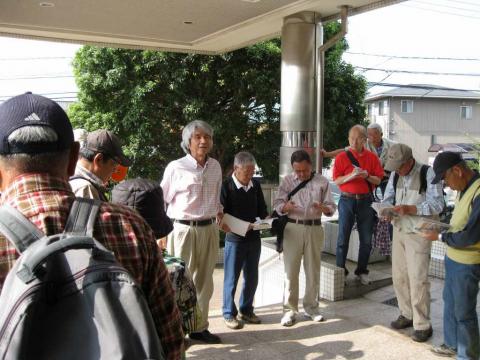 建物のエントランスの雨除けの下に集まった、リュックなどの荷物を持ったたくさんの参加者たちが、手に持った資料を確認している様子の写真