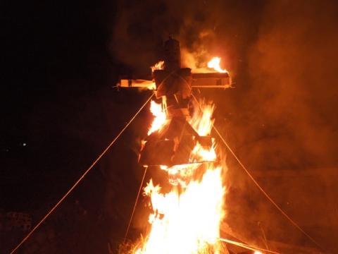 暗闇の中、四方にロープが張られた大きな藁人形が炎を上げて燃えている下大槻百八炬火の様子の写真