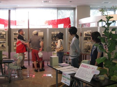 広間で行われている展示会で、来場者たちが壁に飾られた白黒のパネルを熱心に鑑賞しており、手前にはタバコの植物や関連資料が置かれたテーブルがある会場の様子の写真