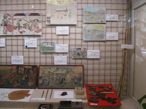 壁に設置したワイヤーネットに複数の絵や説明が掲示され、その下の台にはキセルや煙草入れなどの道具や、額に入った日本画が展示されている展示スペースの写真