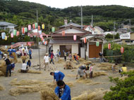 たくさんの提灯が飾られた山あいの集落の広場で、たくさんの人々が、地面に置かれたわらを束ねたり運んだりしている様子の写真