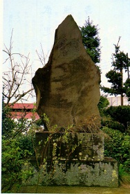 植栽の中、苔が生えた石の台座の上にそびえ立つ、表面に文字のようなものが刻まれた、先端が尖った形をした大きな石碑の写真