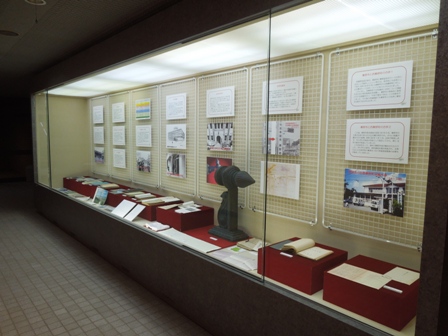 ガラスケース内の壁に複数の説明パネルや写真が掛けられ、赤い台の上に資料などが並べられている展示の様子の写真