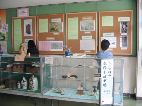 ツボや木箱、草鞋などの道具が陳列された展示ケースが並び、壁のボードに掲示された資料を2人の女性が眺めている、展示会場の様子の写真