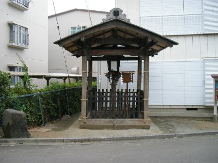 住宅地の一角に建てられた灯籠と、その周りを囲む木製の屋根と柵で囲まれた覆屋の写真