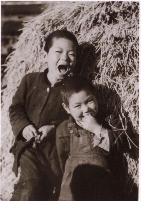 坊主頭の男の子2人が、藁の山にもたれて立ち、手前の小さな男の子は口に指をくわえ、後ろの男の子は口を大きく開けている白黒写真