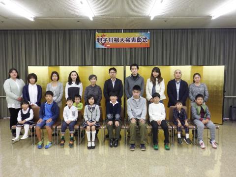 「親子川柳大会表彰式」の横断幕の下に設置した金屏風の前で、前列の椅子に9人の子どもたちが座り、後列に10人の保護者の方たちが並んで立って写した記念写真
