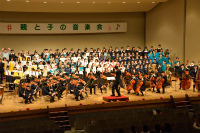 舞台上で指揮者のもと、手前は弦楽器を演奏している生徒たちと後方に白いシャツや青いブレザーを着た合唱団の生徒たちが多数参加して演奏する様子を客席側から捉えた写真