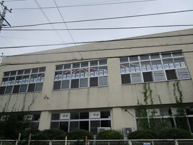 クリーム色の外壁を持つ2階建ての建物で、窓に多数のポスターが掲示され、壁面に蔦が這い、電線が上部を横切る曇天下の西公民館の写真