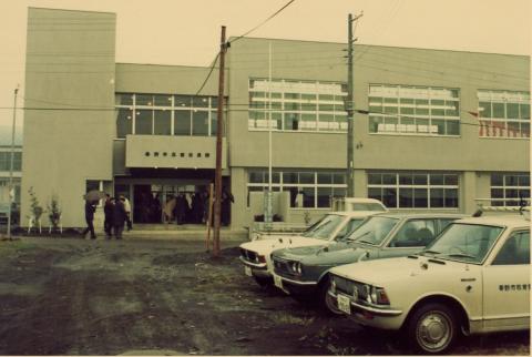 淡い緑色の外壁を持つ2階建ての建物前に複数の白いタクシーが停車し、人々が建物の入口付近に集まっている40年前の西公民館の写真