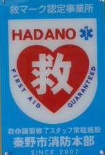 白地に赤いハートマークと救の文字、HADANOロゴが入った秦野市消防本部の救マーク認定事業所ステッカーの写真
