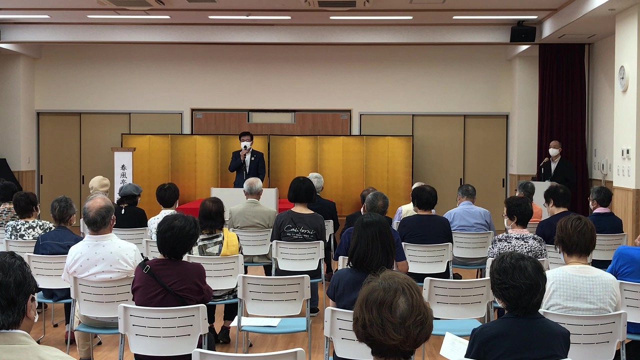 金屏風と赤い布の演台を背景にスーツ姿の高橋市長が挨拶し、白い椅子に座った多数の参加者が聞いている室内での公式行事の写真