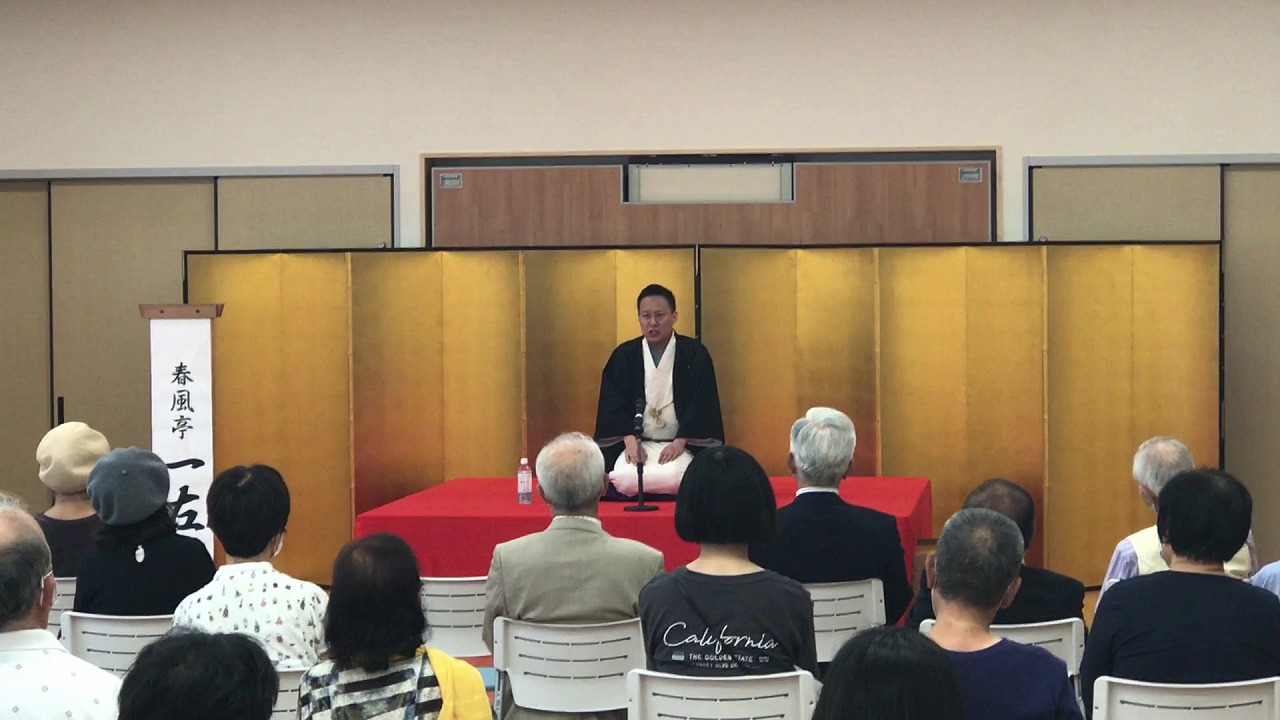 金屏風を背景に赤い布の演台で黒い着物姿の男性が座り、春風亭一左の文字が入った立て看板の前で白い椅子に座った観客が落語を聞いている写真