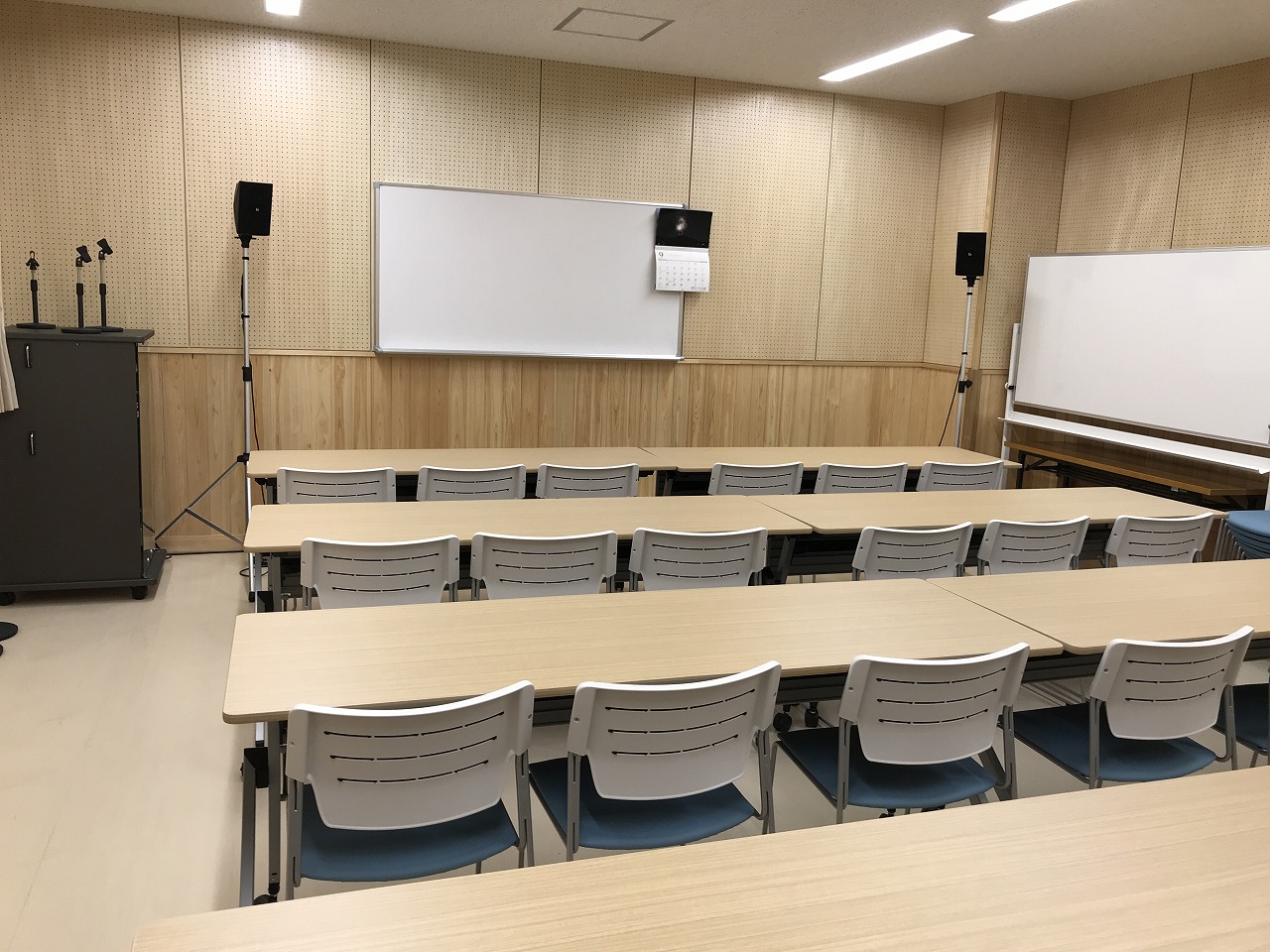ホワイトボードやスピーカーが設置され、木目調の長机、白い椅子が整然と並んでいる明るい研修室の写真
