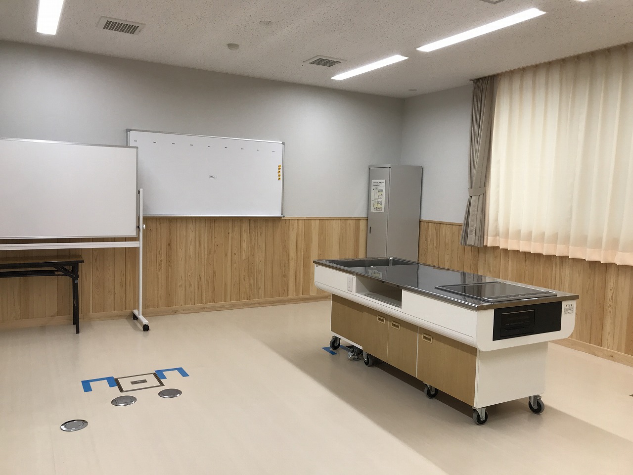 木目調の壁と白い床の部屋に、可動式の調理台と2枚のホワイトボードが設置されている、明るい雰囲気の集会室Bの写真