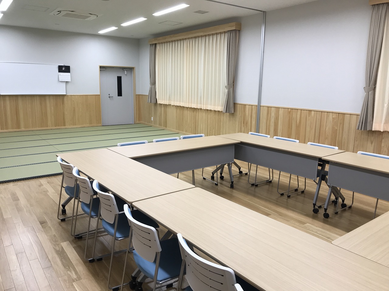 和室とフローリングのスペースが共存する部屋で、木目調のテーブルがロの字型に配置され、青と白の椅子が並べられた集会室Dの写真