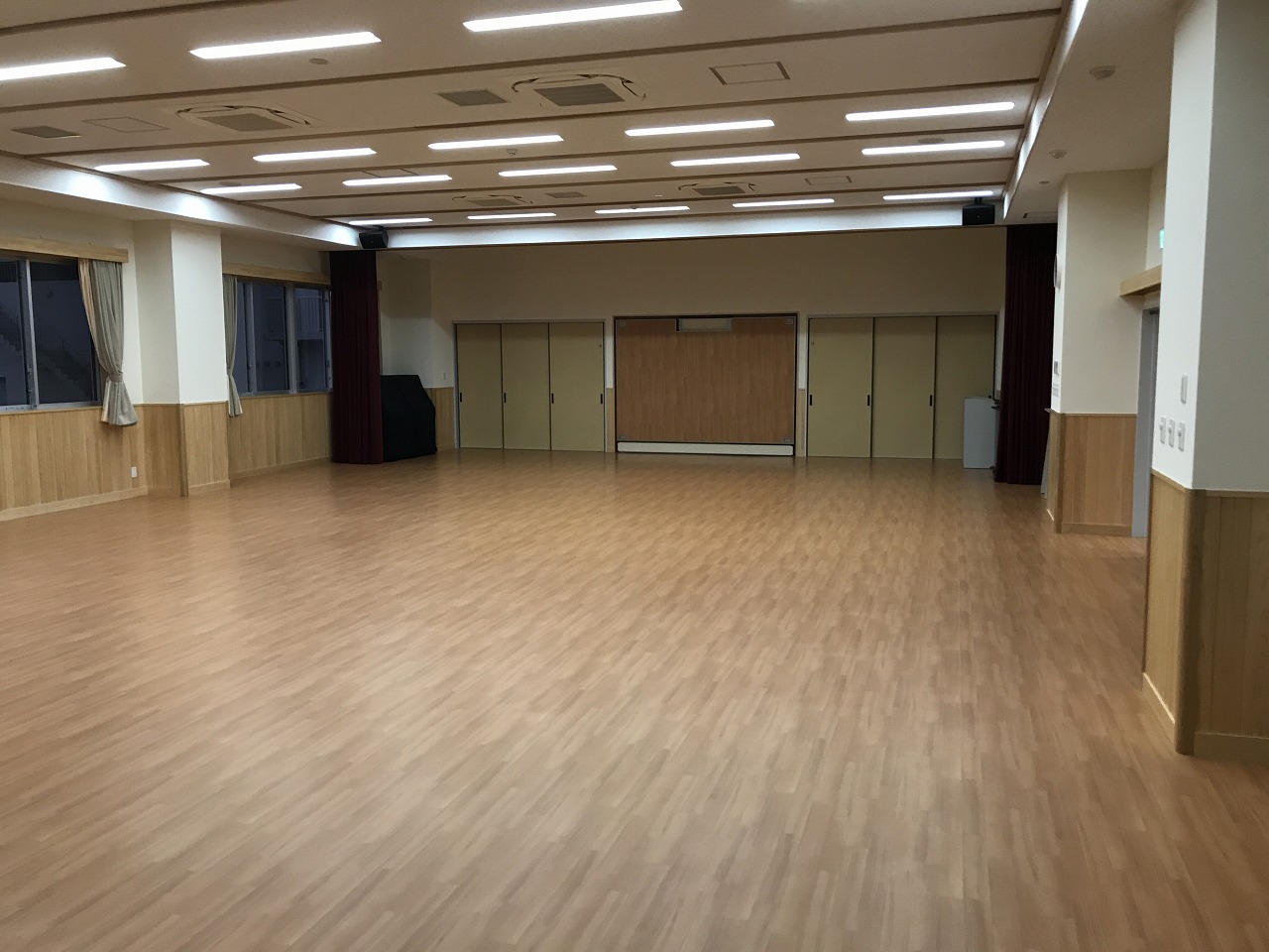 フローリングの床と木目調の壁が特徴的で、正面に襖や可動式パネルがある多目的ホールの写真