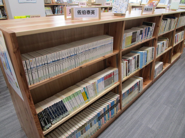 木製の本棚に佐伯泰英と書かれた白い札が立ち、下段に黒や白の背表紙の文庫本がシリーズで整然と並ぶ図書館の書架の写真