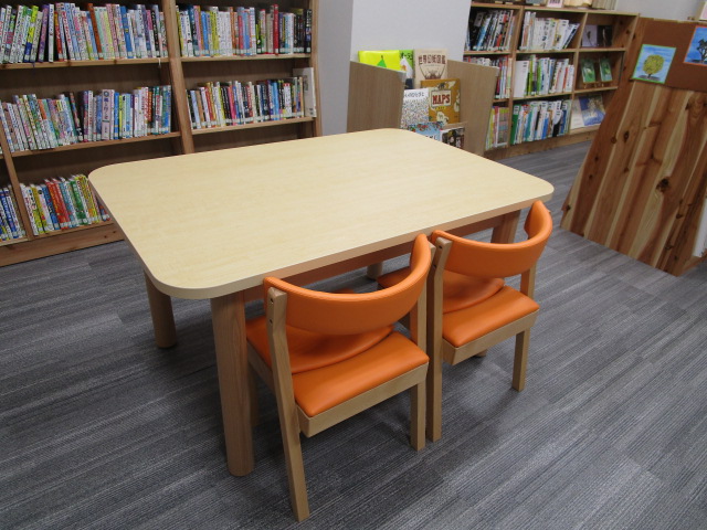木製の本棚が並ぶ図書館内に、白い天板の木製テーブルとオレンジ色の背もたれ付き木製椅子2脚が置かれた閲覧スペースの写真