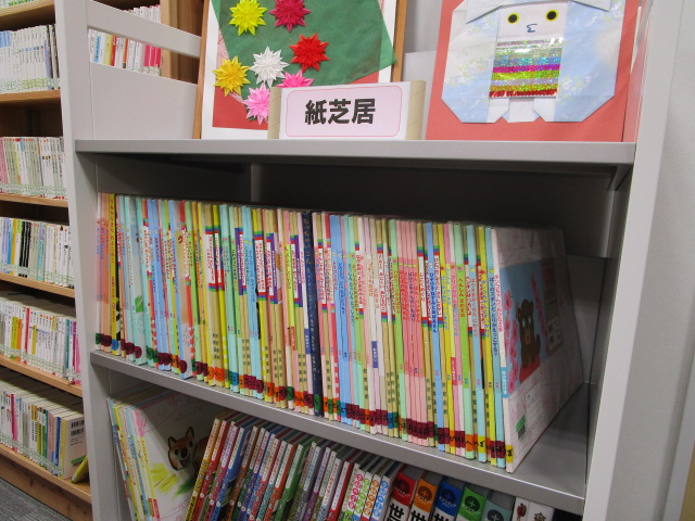 白い本棚の上部に紙芝居と書かれた札と花の飾りが置かれ、棚にカラフルな背表紙の紙芝居作品が整然と並ぶ図書館の紙芝居コーナーの写真
