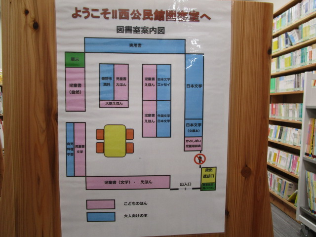 児童書や実用書、日本文学、外国文学など、色分けされた本の配置がわかる西公民館図書室の案内図が木製の本棚の横に掲示されている写真