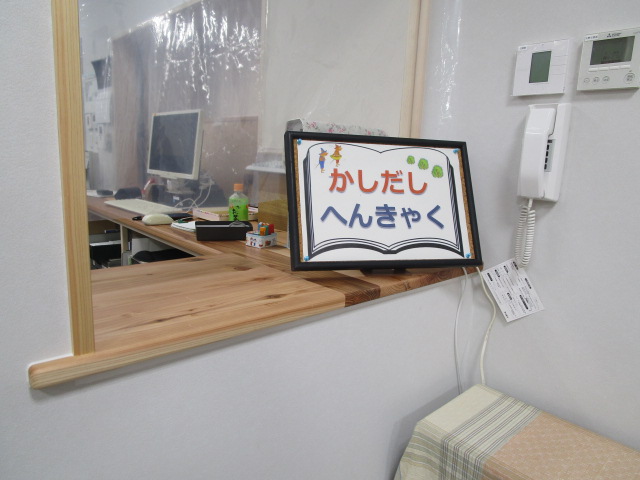 感染症対策の透明なビニールカーテンが吊るされた木製のカウンターの上に、開いた本のイラストとともに平仮名で「かしだし」「へんきゃく」と書かれた案内板が設置されている窓口の写真