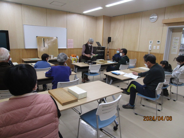 教室で数人の参加者が机に座り、前方の講師が資料を見せながら説明している様子を写した写真