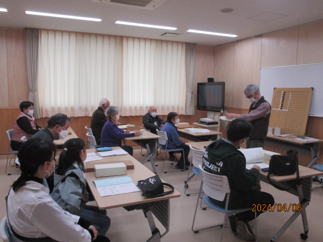 教室で複数の参加者が机に座り、講師が前方で資料を使って説明している様子を写した、講座説明する様子の写真