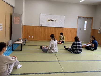 畳敷きの部屋で数人の保護者の前に赤ちゃんを寝かせ、壁には「赤ちゃんわらべうた」の看板が掛かっている、赤ちゃんわらべうた教室の写真