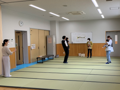 畳敷きの部屋で数人の保護者が立って赤ちゃんを抱いており、壁には「赤ちゃんわらべうた」の看板が掛かっている、赤ちゃんわらべうた教室の写真