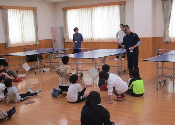 卓球教室で、指導者が座っている子供たちにラケットの持ち方や打ち方などの技術を教えている様子を写した、児童向け卓球指導の写真